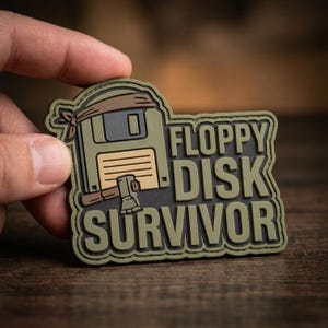 May include: A tactical morale patch in olive green, tan, and black featuring a stylized floppy disk wearing a headband with an axe resting against it. The text FLOPPY DISK SURVIVOR is embossed in large, bold, raised lettering across the right side.