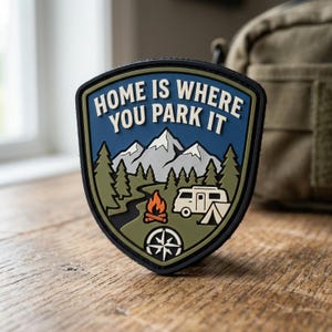 May include: A shield-shaped patch with a black border featuring the text "HOME IS WHERE YOU PARK IT" in white. The design depicts a blue sky, white snow-capped mountains, green pine trees, a winding river, a campfire, a camper van, and a compass.