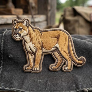 May include: An embroidered patch featuring a mountain lion in profile, rendered in shades of tan, brown, and cream. The cougar is depicted standing with its head turned slightly toward the viewer, outlined with a dark brown stitched border.