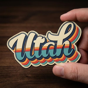 May include: A retro-style sticker with the word "Utah" in a layered, cursive font. The design features a cream-colored outline with blue, orange, and yellow stripes, evoking a vintage aesthetic. The sticker is held against a dark wooden background.