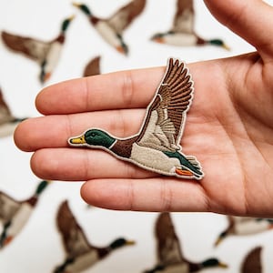 May include: Embroidered patch of a flying mallard duck. The patch features detailed stitching in shades of brown, green, and beige, with a yellow beak and orange feet. The duck is shown mid-flight with its wings spread. Other duck patches are in the background.