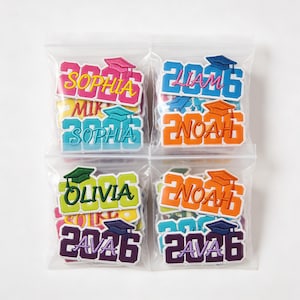 May include: Four clear plastic bags each containing graduation-themed patches. Each patch features the year "2026" and a name, such as "Sophia" or "Liam", with a graduation cap. The patches are in various colors, including pink, blue, and orange.