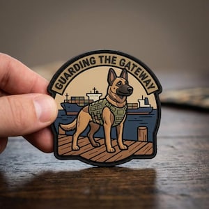May include: A circular morale patch featuring a tan German Shepherd wearing an olive green tactical vest, standing on a wooden dock. In the background, a cargo ship sits on blue water. The text GUARDING THE GATEWAY is printed in black.