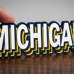 May include: A navy blue and yellow Michigan state wordmark sticker. The word "MICHIGAN" is in white, with a navy blue border and yellow accents. The sticker is held by a hand.
