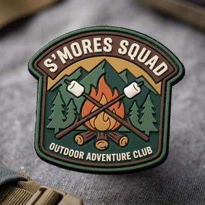May include: A shield-shaped PVC patch featuring a campfire with two crossed sticks holding marshmallows. The design includes green mountains, pine trees, and the text S'MORES SQUAD and OUTDOOR ADVENTURE CLUB in cream lettering against a dark green background.