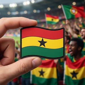 May include: A rectangular patch featuring the flag of Ghana, held in front of a crowd. The flag has horizontal stripes of red, yellow, and green, with a black star in the center. The background shows fans waving flags.