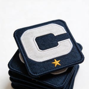 May include: A stack of navy blue square patches with a white letter "C" and a small yellow star. The patches have a stitched border and a soft, textured appearance. The patches are stacked on top of each other.