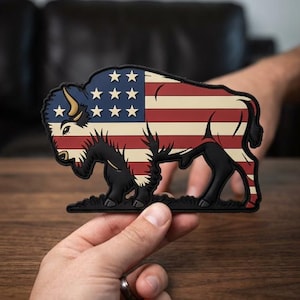 May include: A bison-shaped patch featuring the American flag design. The patch has a blue section with white stars, and red and cream stripes. The bison is black with cream-colored horns and a black base. The patch is held by a person.
