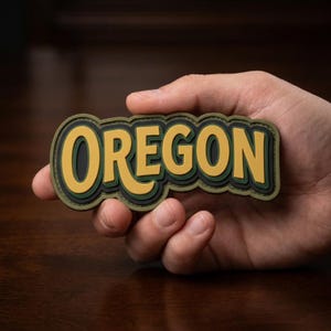 May include: A layered patch featuring the word "OREGON" in bold, yellow letters. The lettering is outlined in black and green, creating a 3D effect. The patch is held in a hand against a blurred background.