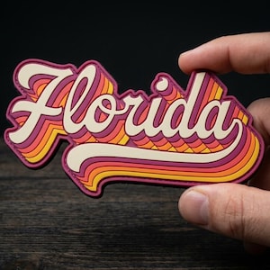 May include: A colorful Florida-themed sticker with a retro design. The word "Florida" is written in a layered, stylized font with shades of pink, orange, yellow, and white. The sticker is held against a dark background.