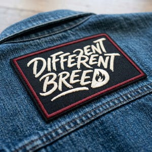 May include: A rectangular black patch with the words "DIFFERENT BREED" in white script. The patch has a red border and a small flame graphic. It is placed on a blue denim jacket.
