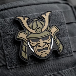 May include: A detailed patch featuring a samurai helmet design. The patch has a black border with tan, olive green, and white accents. The patch is attached to a dark gray fabric.