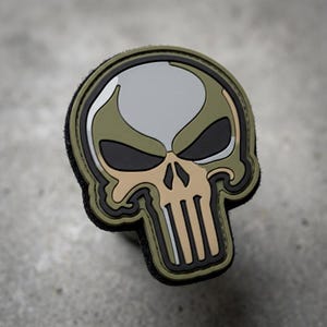 May include: A patch featuring a skull design with a gray, tan, black, and olive green color scheme. The patch has a black border and a hook-and-loop backing.