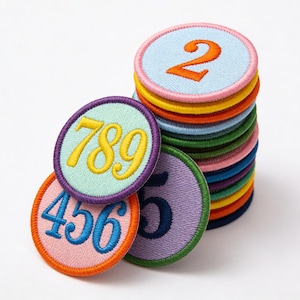 May include: A stack of colorful, embroidered number patches. Each patch features a different number from 2 to 9, with a variety of colors including blue, orange, pink, and green. The patches are round with a contrasting border.