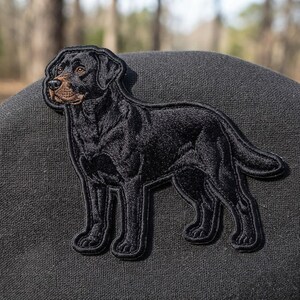 May include: An embroidered patch featuring a black dog with a brown muzzle and ears, standing in profile. The detailed stitching captures the dog's fur texture and facial features, with a black border outlining the entire shape of the animal.