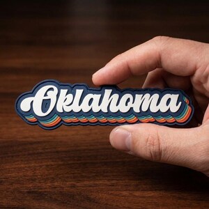 May include: A retro-style Oklahoma state souvenir. The word "Oklahoma" is in white, outlined in navy blue, with a wavy design in shades of blue, orange, and yellow. The item is held in a hand.
