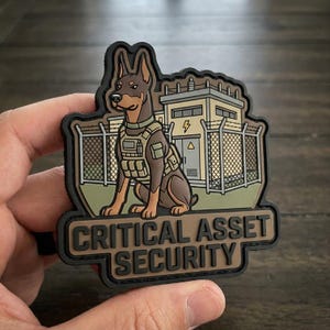 May include: A tactical morale patch featuring a brown Doberman dog wearing a tan tactical vest, positioned in front of a fenced industrial building with a lightning bolt symbol. The text CRITICAL ASSET SECURITY is embossed in black at the bottom.