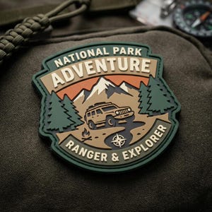 May include: A shield-shaped rubber patch featuring the text NATIONAL PARK ADVENTURE and RANGER & EXPLORER. The design includes a tan off-road vehicle, dark green pine trees, a small campfire, a compass rose, and snow-capped mountains in shades of tan and orange.
