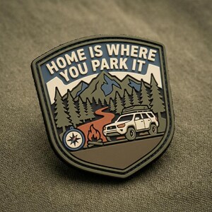 May include: A shield-shaped morale patch featuring a white SUV with a roof rack, a campfire, a compass, and a mountain forest landscape. The text HOME IS WHERE YOU PARK IT is printed in white at the top against a blue sky background.