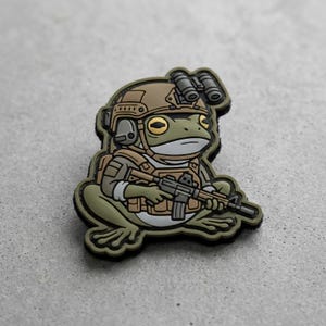 May include: A patch featuring a cartoon frog in military gear, including a helmet, vest, and rifle. The frog is primarily green with tan and black accents. The patch has a black border and is set against a neutral background.
