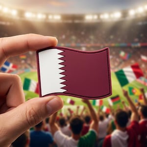 May include: A maroon and white flag of Qatar is held up in front of a crowd at a sporting event. The flag has a serrated edge on the white portion. The background is blurred, showing spectators and other flags.