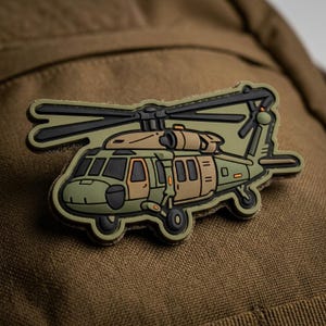 May include: A green and tan helicopter patch with black details. The patch features a detailed illustration of a helicopter with its rotors and tail. The patch is likely made of rubber or a similar material and is designed to be attached to clothing or gear.