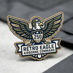 May include: A patch featuring a stylized eagle with outstretched wings, wearing a pilot's helmet. The design includes the text "RETRO EAGLE AIRBORNE COMMAND" and the number "250". The patch is in shades of blue, tan, and white.
