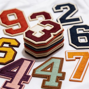 May include: A collection of embroidered number patches in various colors, including red, navy, yellow, and purple. The patches have a classic varsity style with a white border and are stacked and scattered on a white surface.