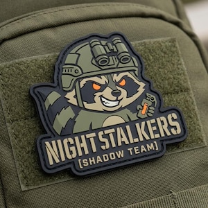 May include: A patch featuring a cartoon raccoon wearing a military helmet and holding a grenade. The patch has the words "NIGHT STALKERS" and "[SHADOW TEAM]" in a rectangular border. The patch is black, tan, and olive green.