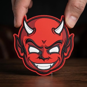 May include: A red devil face patch with white horns, eyes, and teeth, and black outlines. The patch is held between two fingers against a dark background. The devil has a wide, toothy grin.