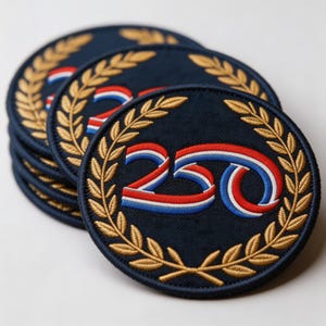 May include: A stack of navy blue circular patches. Each patch features the number "250" in red, white, and blue, encircled by a gold laurel wreath. The patches are embroidered with a textured appearance, suggesting a high-quality finish.
