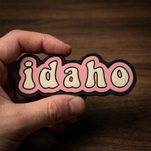 May include: A pink and white Idaho patch with a black border. The word "Idaho" is written in a retro font. The patch is held in a person's hand against a dark wood background.