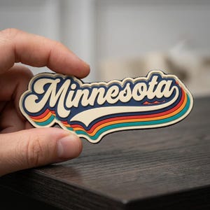 May include: A retro-style magnet with the word "Minnesota" in a cream-colored, cursive font. The text is layered over a blue background with colorful stripes of orange, red, and teal. The magnet is held against a dark wooden surface.