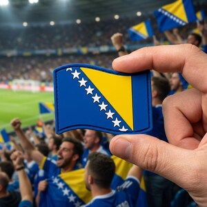 May include: A blue, yellow, and white flag of Bosnia and Herzegovina is held up in front of a crowd of cheering fans. The flag has a blue background with a yellow triangle and white stars.