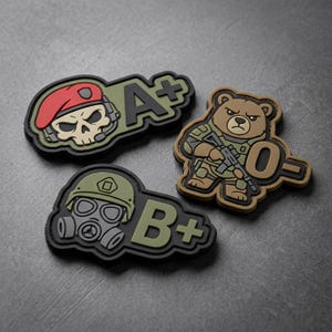 May include: Three tactical patches with military-themed designs. One patch features a skull wearing a red beret and headset with the text "A+". Another shows a bear in camouflage holding a rifle with "O-". The third patch displays a gas mask and helmet with "B+".