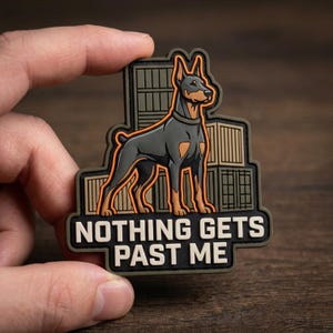 May include: A rectangular rubber patch featuring a Doberman dog standing in front of stacked shipping containers. The design includes a bright orange outline around the dog and the text "NOTHING GETS PAST ME" in bold, light-colored capital letters.
