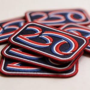 May include: A stack of embroidered patches with the number "250" in red, white, and blue stitching on a navy blue background. The patches have a rectangular shape with a red border. The patches are stacked on top of each other.