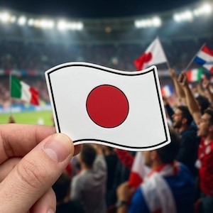 May include: A white flag with a red circle in the center, representing the Japanese flag, is held up in front of a crowd. Other flags are visible in the background, suggesting a sporting event or international gathering.
