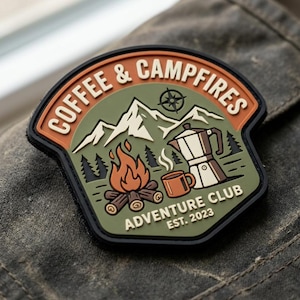 May include: A shield-shaped patch featuring an olive green background with a mountain range, a compass, a campfire, a coffee mug, and a moka pot. The text reads COFFEE & CAMPFIRES ADVENTURE CLUB EST. 2023 in cream-colored lettering on an orange border.