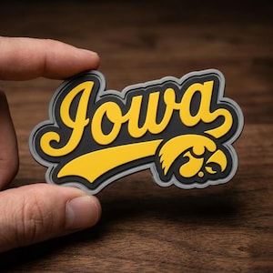 May include: A black, yellow, and gray Iowa Hawkeyes logo. The word "Iowa" is in yellow script, with a yellow hawk head logo. The logo has a gray border and is held by a hand.