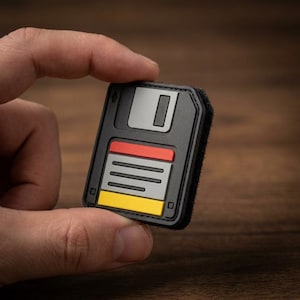 May include: A square, black, rubberized patch shaped like a vintage floppy disk. The design features a grey sliding metal shutter, a red horizontal bar, a grey label area with horizontal lines, and a yellow rectangular tab at the bottom edge.