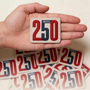 May include: A rectangular patch with the word "AMERICA" above the number "250". The "2" and "0" are red, and the "5" is navy blue. The patch is beige with a stitched border. Several similar patches are scattered below.