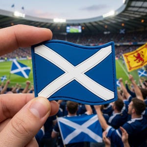 May include: A blue and white patch featuring the Scottish flag, held up in front of a crowd of people. The flag has a white X on a blue background. Other Scottish flags are visible in the background, along with a yellow flag.