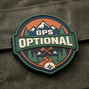 May include: A circular rubber patch featuring a mountain landscape with pine trees, a winding path, a compass, and crossed axes. The text GPS OPTIONAL is displayed in cream-colored lettering against a dark blue and green background with orange accents.
