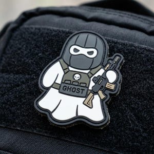 May include: A black and white rubber morale patch featuring a cartoon ghost character wearing a dark grey tactical balaclava, a green plate carrier vest with a skull emblem, and the word GHOST printed on the front while holding a rifle.