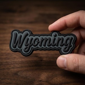 May include: A black and gray patch with the word "Wyoming" in a stylized font. The patch is held by a hand against a dark wood background. The lettering is raised, creating a 3D effect.