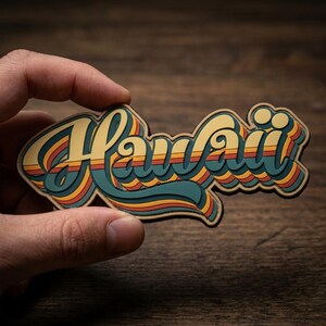 May include: A retro-style sticker with the word "Hawaii" in a layered design. The lettering is yellow with blue, orange, and red stripes, and a brown border. The sticker is held against a wooden background.
