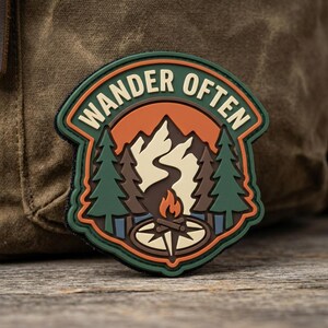 May include: A circular patch with a dark green border featuring the text WANDER OFTEN in cream letters. The design depicts a campfire, a compass, two pine trees, and a mountain range in shades of orange, brown, cream, and green against a dark background.