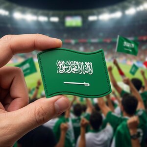 May include: A green patch featuring the Saudi Arabian flag, held in a hand. The flag design includes white Arabic script and a white sword on a green background. The background shows a crowd of people, some holding similar flags.