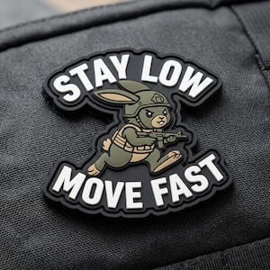 May include: A black and white patch with the words "STAY LOW" and "MOVE FAST". The patch features a cartoon rabbit wearing a helmet and holding a rifle. The rabbit is colored in shades of green and brown.